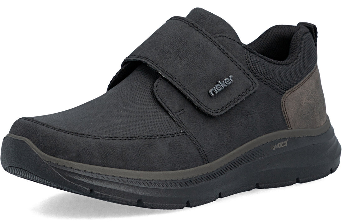 A black slip-on shoe with a hook-and-loop strap closure and a textured sole, shown from a three-quarters angle on a white background.
